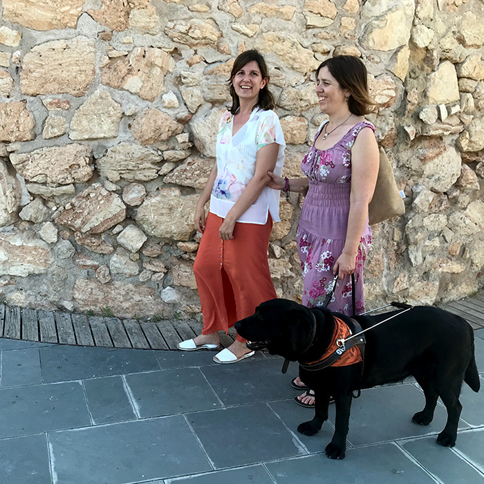 La consultora Andrea Granell acompanyant una dona amb ceguesa i el seu gos pigall. Al darrera, torre de pedra d’estructura circular, és la Torre Blava-Espai Guinovart situada al Passeig Marítim de Vilanova i la Geltrú.