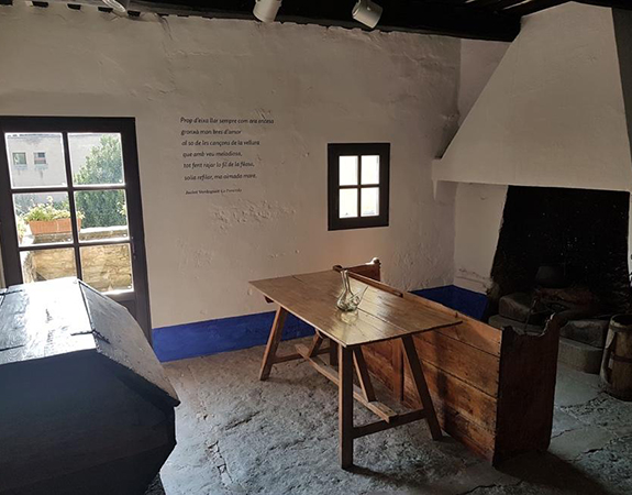Cuina de casa de pagès del segle dinou amb una porta i una finestra petita, una taula de fusta amb un porró al centre i una llar de foc.